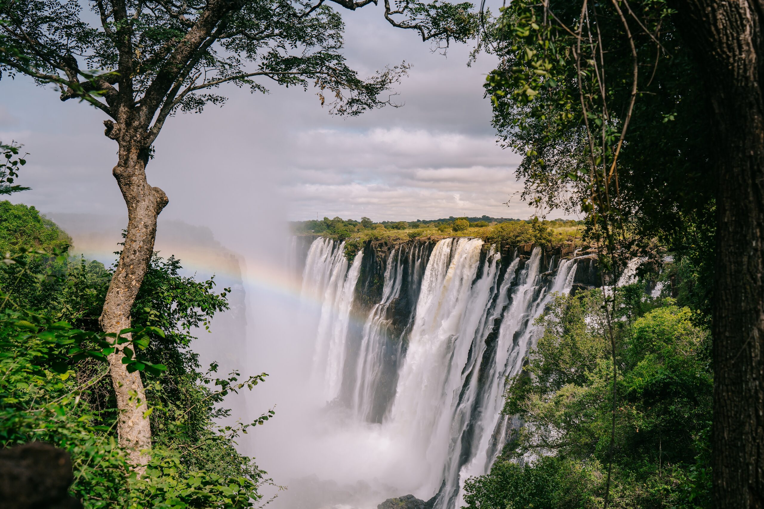 Klassiek uitzicht op de Victoria Falls vanaf de Zambiaanse kant, met regenboog in de mist en dramatische wolken – Christoffel Travel.